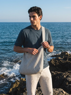 A man standing on a rocky beach wearing a navy blue t-shirt and light beige linen pants, carrying a large cream and navy striped canvas shoulder bag.