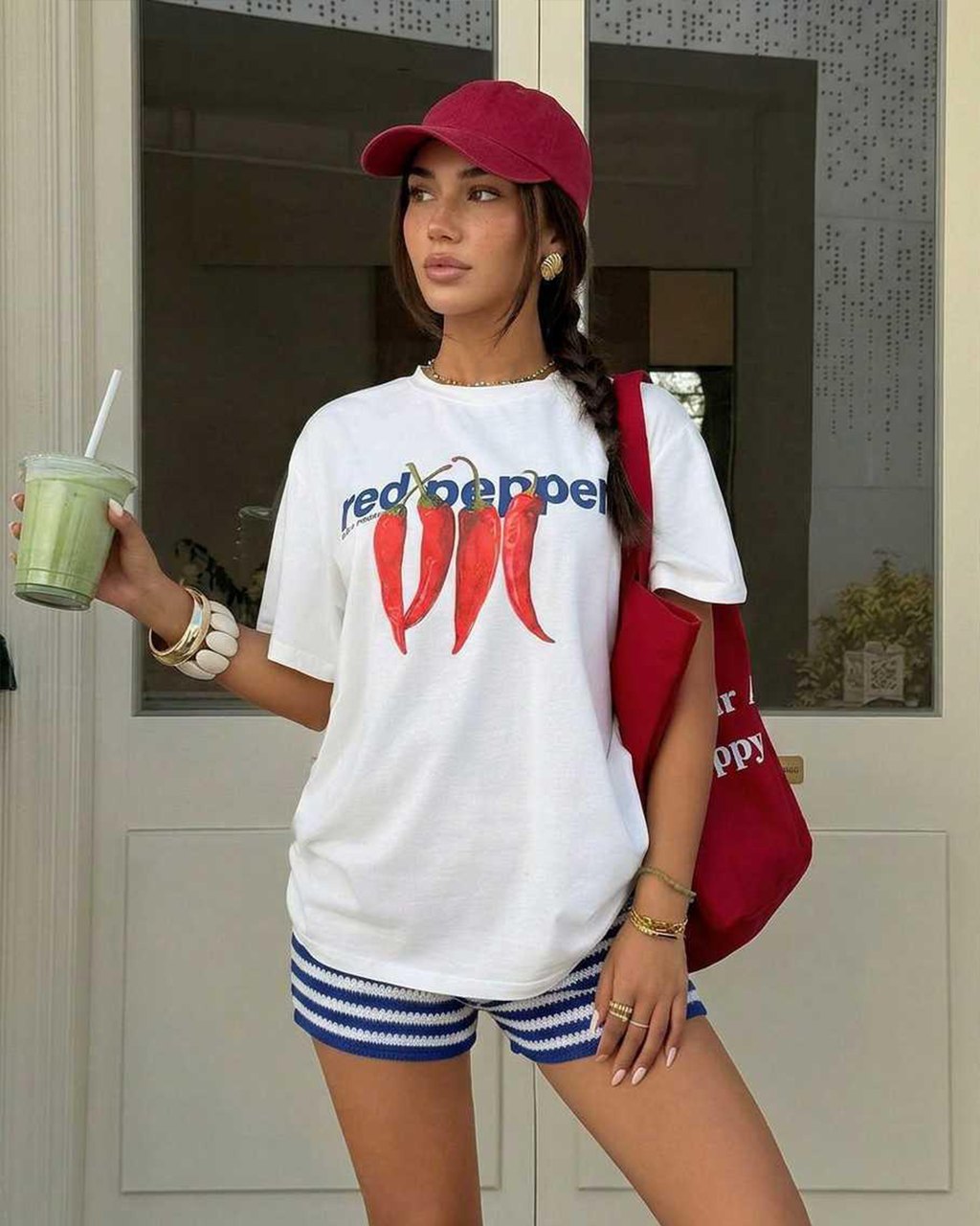 Person wearing a white t-shirt with red and blue graphics, holding a drink and a red bag.
