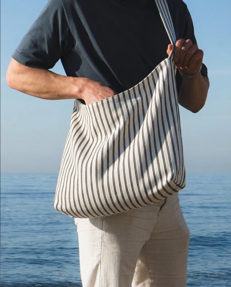 A person standing by the ocean wearing a black t-shirt and light beige pants, carrying a large slouchy shoulder bag with vertical black stripes on a cream fabric background.